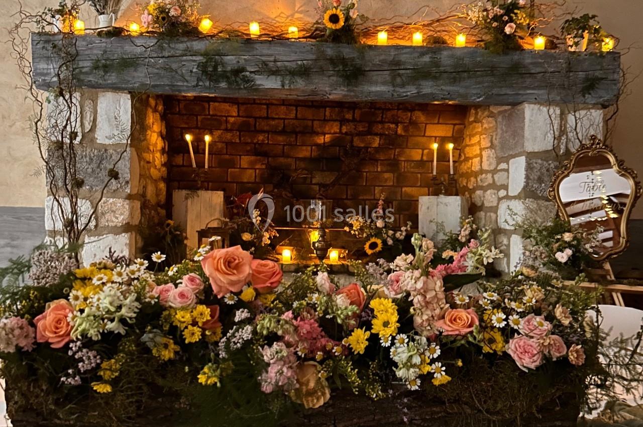 Cheminée décorée de fleurs colorées, bougies allumées et guirlande lumineuse dans un cadre rustique.