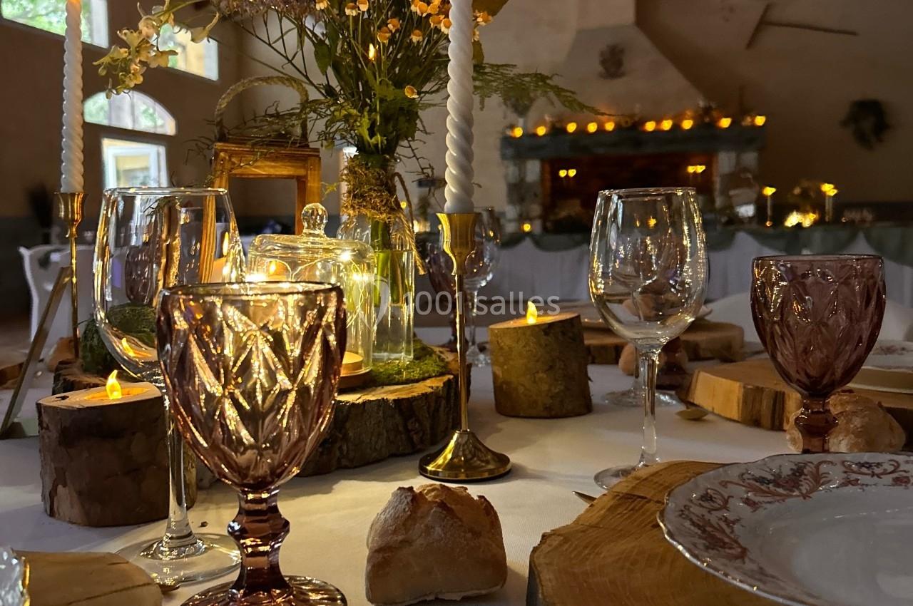 Table décorée pour un dîner, avec bougies allumées, verres colorés, rondins de bois et fleurs dans une salle chaleureuse.