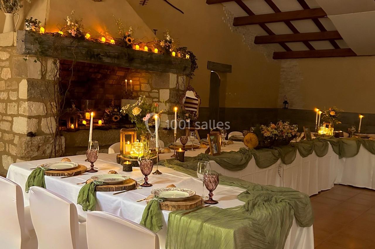 Salle décorée pour un repas, avec tables dressées, nappes blanches, voilages verts et cheminée ornée de bougies.
