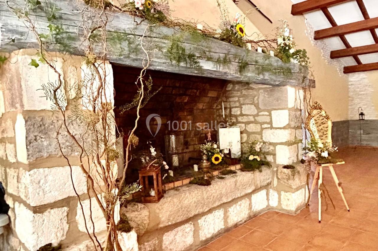 Cheminée en pierre décorée de fleurs et branchages dans une pièce avec plafond en poutres apparentes.