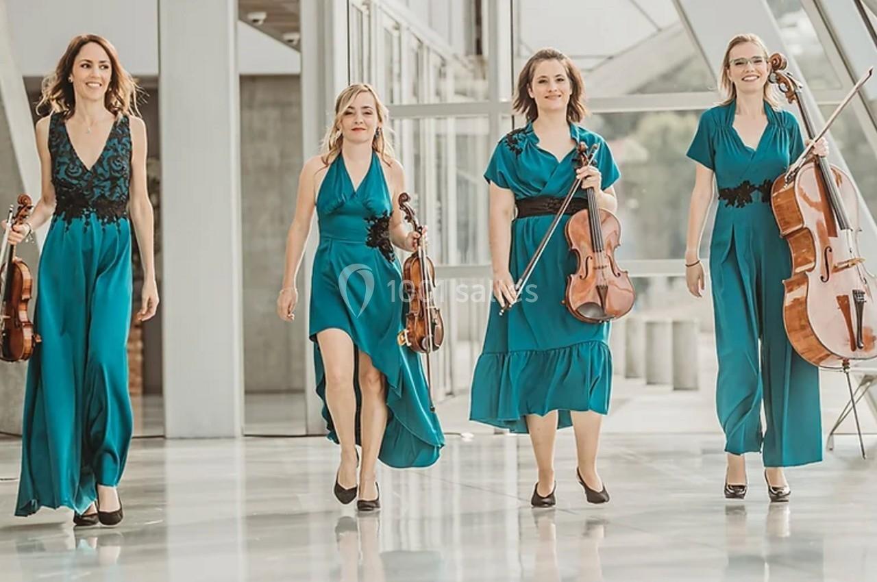 Quatre musiciennes en robes turquoise portant des instruments à cordes marchent dans un espace lumineux et moderne.