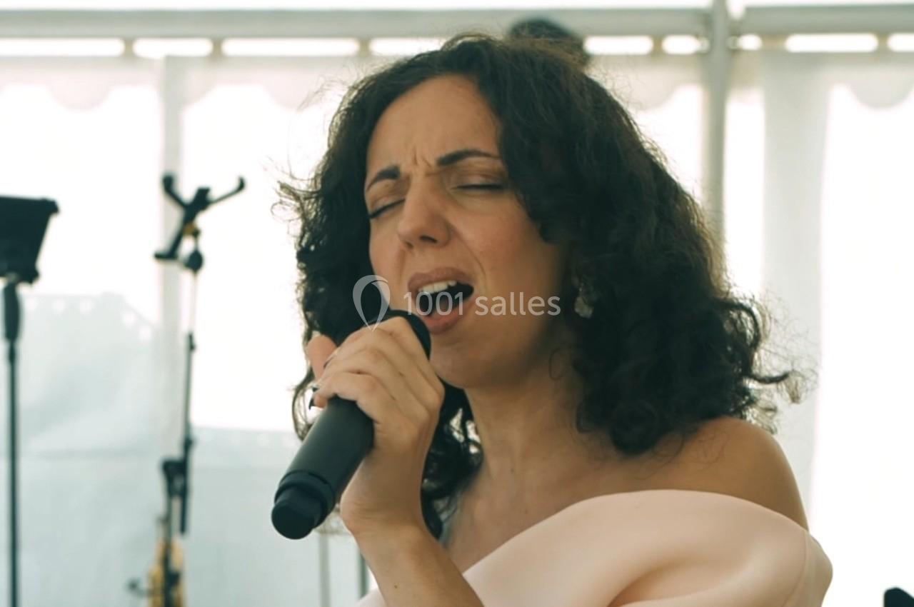 Femme chantant avec un micro à la main, en intérieur lumineux avec des équipements musicaux en arrière-plan.