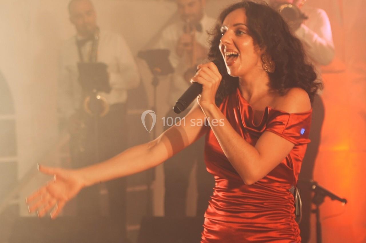 Une chanteuse en robe rouge interprète sur scène, accompagnée de musiciens en arrière-plan.