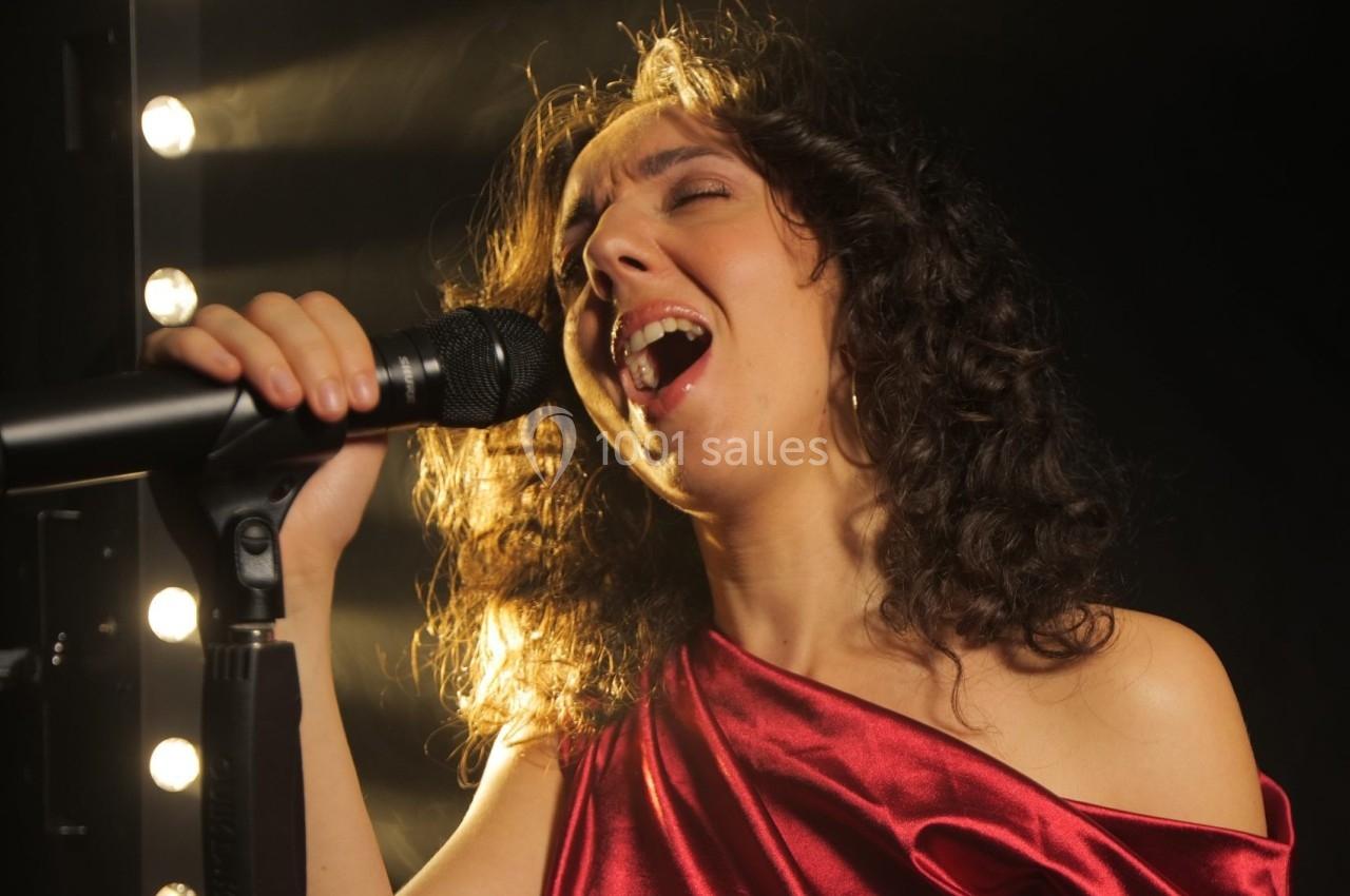 Femme chantant avec passion dans un micro, éclairée par des projecteurs sur un fond sombre.