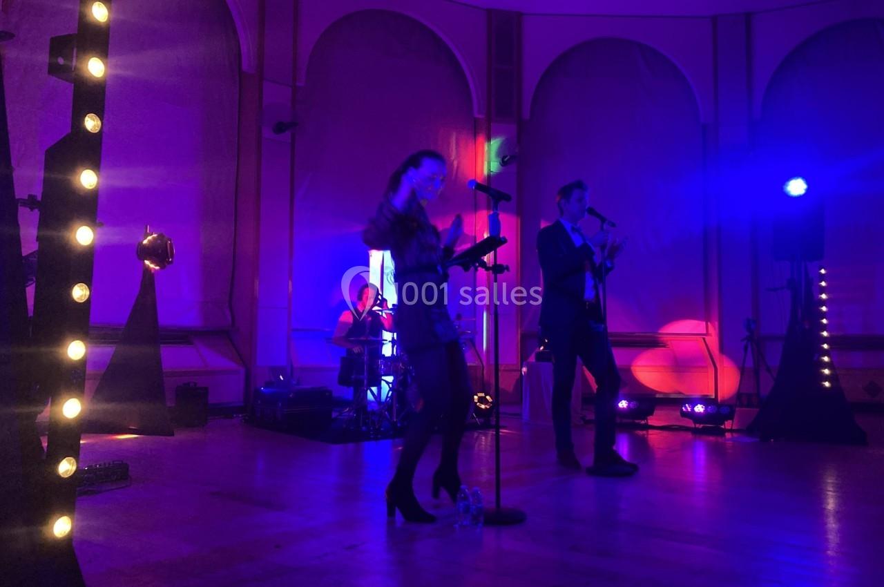 Groupe de musiciens jouant sur une scène éclairée de lumières colorées dans une salle avec de grandes fenêtres.