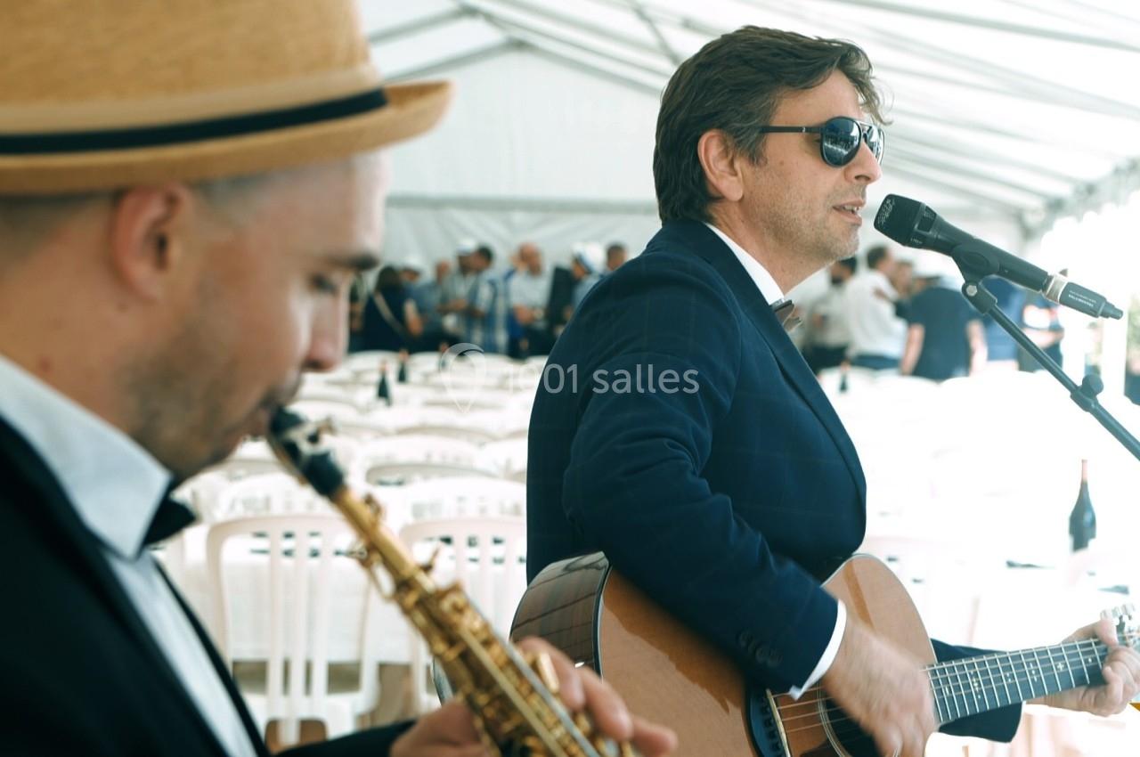 Deux musiciens jouent de la guitare et du saxophone sous une tente blanche lors d'un événement en plein air.