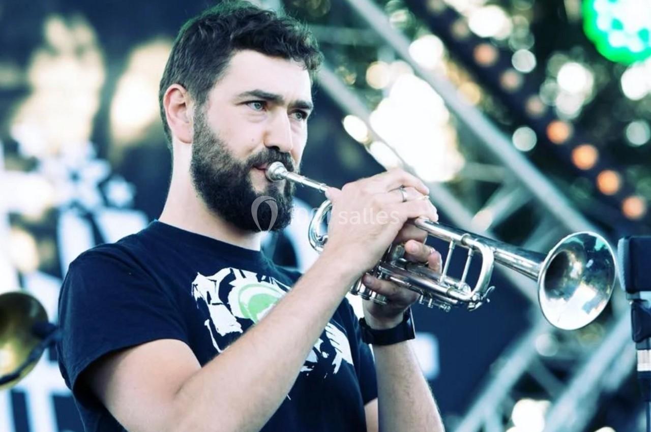 Un homme barbu joue de la trompette sur une scène en plein air, éclairée par des lumières colorées.