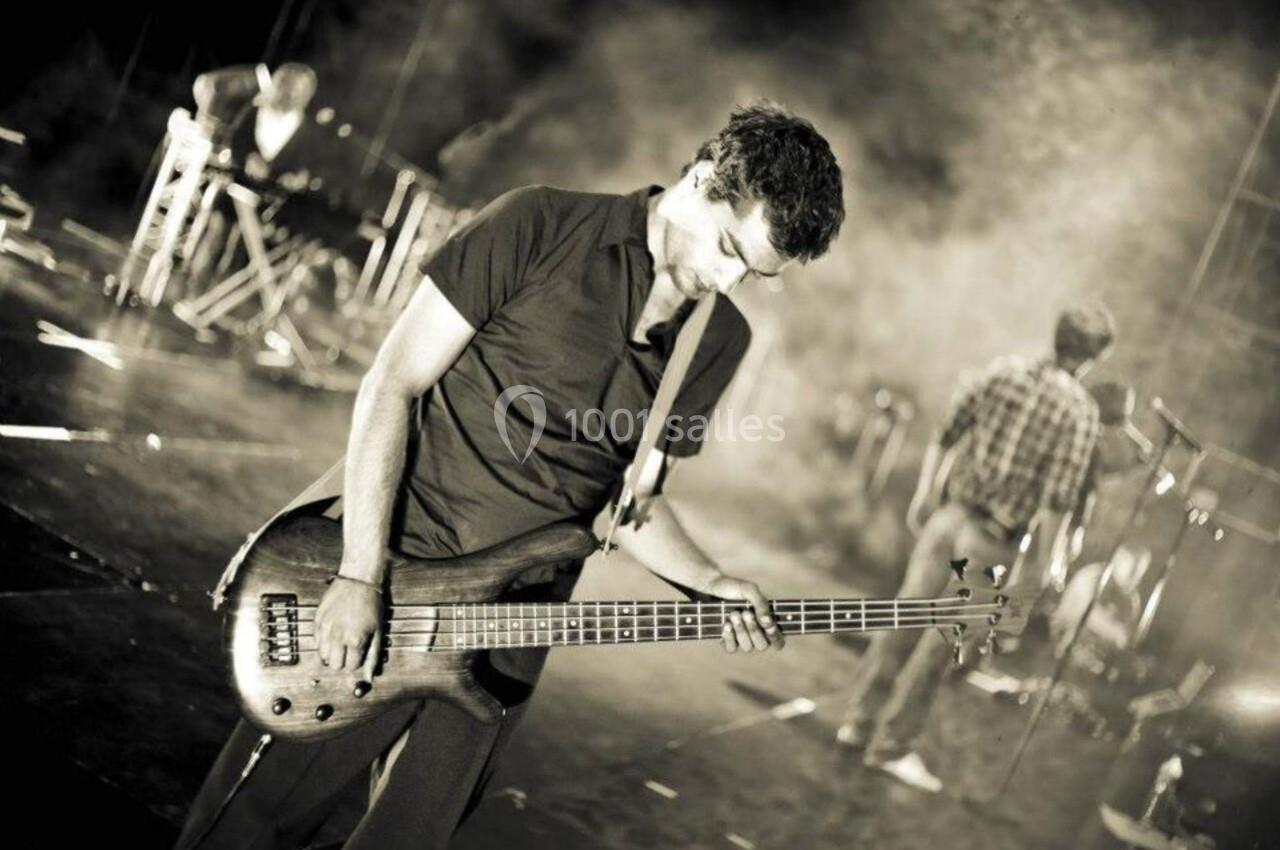 Un musicien joue de la guitare basse sur une scène, éclairée par des projecteurs, avec d'autres musiciens en arrière-plan.