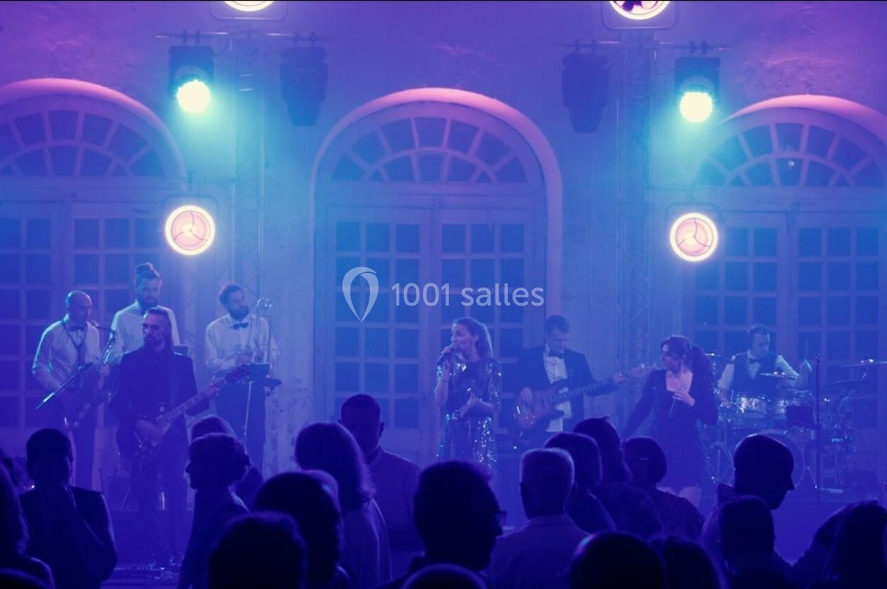 Groupe de musiciens jouant sur scène devant un public, éclairé par des lumières violettes et bleues.