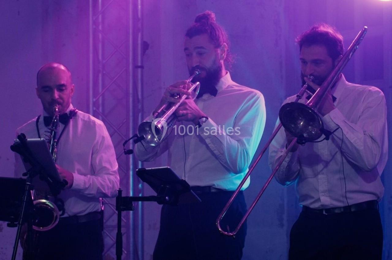 Trois musiciens jouent du saxophone, de la trompette et du trombone sur une scène éclairée par des lumières violettes.
