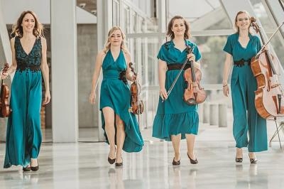Quatre musiciennes en robes turquoise portant des instruments à cordes marchent dans un espace lumineux et moderne.