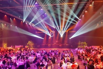 Salle de réception illuminée avec des projecteurs colorés, tables rondes occupées par des convives lors d'un événement.