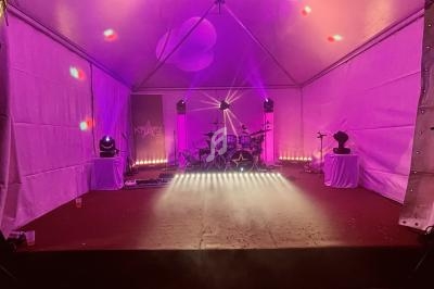 Scène sous chapiteau éclairée de lumières roses et violettes, avec une batterie et des équipements audio.