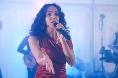 Femme chantant sur scène avec un micro, accompagnée de musiciens en arrière-plan dans une ambiance lumineuse.