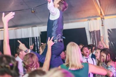 Un homme en costume est porté par des invités dans une ambiance festive sous une tente éclairée.
