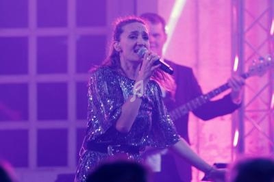 Une chanteuse en robe à paillettes interprète sur scène, éclairée par des lumières violettes, accompagnée de musiciens.