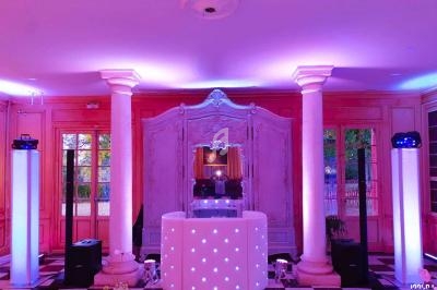 Salle élégante avec colonnes blanches, éclairage rose et violet, et une console DJ lumineuse au centre.