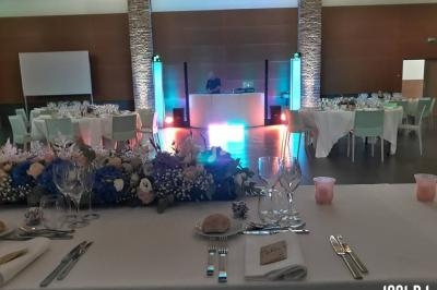 Salle de réception décorée avec des tables dressées, un bouquet floral au premier plan et un DJ en arrière-plan.