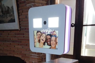 Borne photo avec écran affichant un groupe de personnes souriantes, placée dans une pièce lumineuse avec murs en briques.