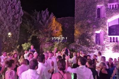 Groupe de personnes dansant en soirée en plein air devant un bâtiment couvert de végétation, éclairé par des lumières…