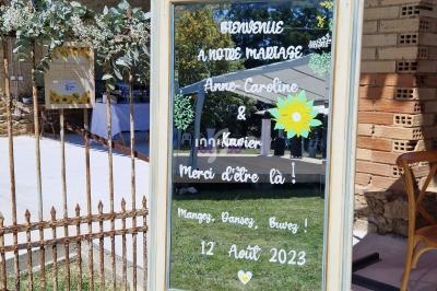 Panneau de bienvenue pour un mariage, écrit à la main sur un miroir encadré, avec décor floral et fond extérieur ensoleillé.