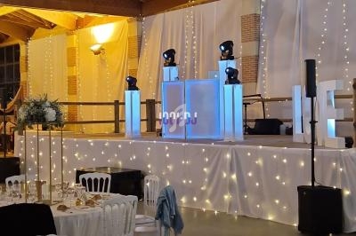 Salle de réception décorée avec des guirlandes lumineuses, une scène équipée de matériel audio et des tables dressées.