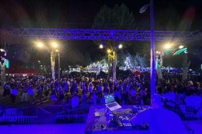 Vue nocturne d'une foule rassemblée pour un événement en plein air, avec un DJ au premier plan et des lumières colorées.