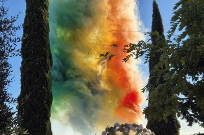 Fumée colorée s'élevant dans le ciel lors d'une cérémonie en plein air entourée d'arbres et de spectateurs assis.