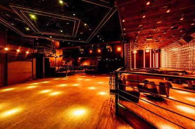 Salle de club vide avec éclairage tamisé, tables rondes, banquettes et escalier menant à une piste de danse.
