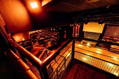 Salle de club vide avec éclairage tamisé, tables rondes, banquettes et escalier menant à une piste de danse.