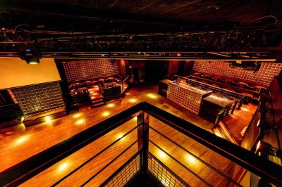 Salle de club vide avec éclairage tamisé, tables rondes, banquettes et escalier menant à une piste de danse.