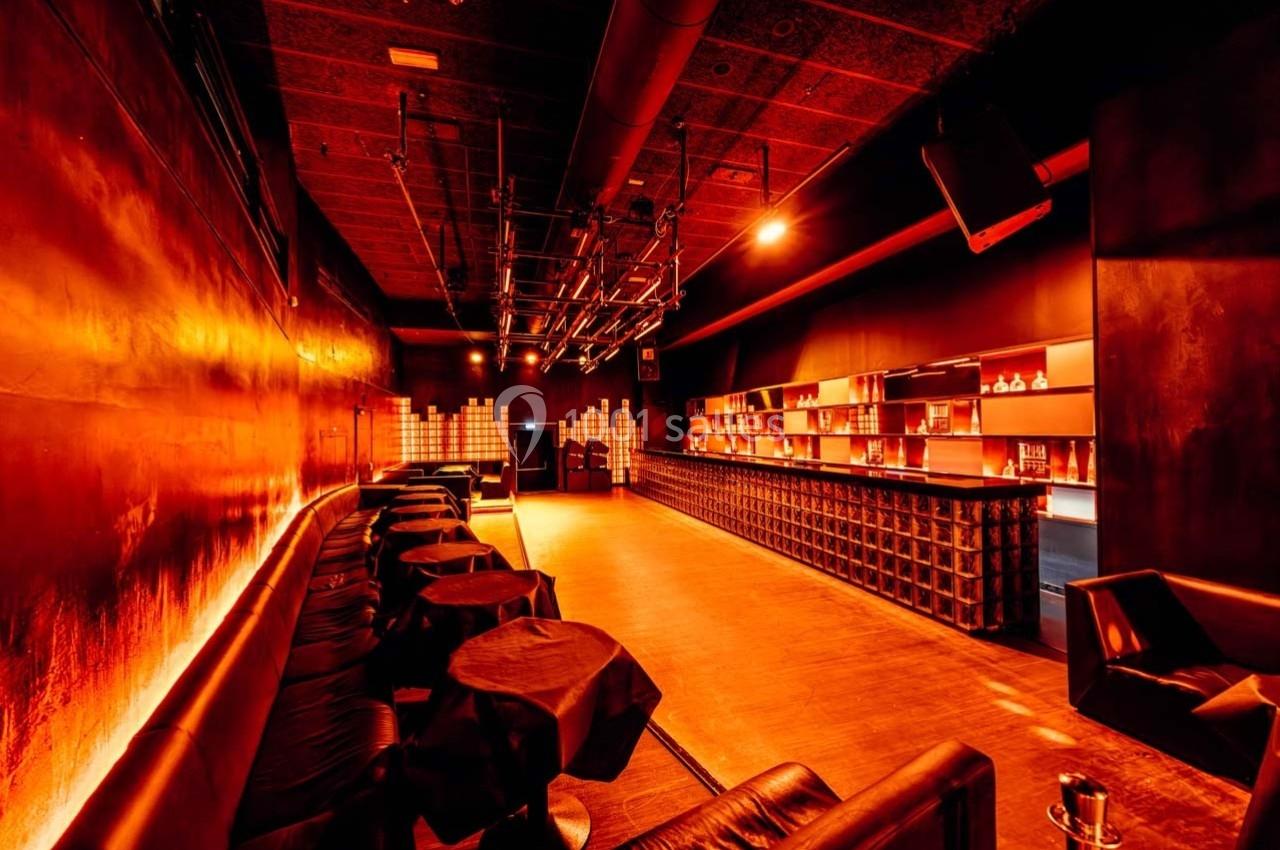 Salle de bar moderne avec éclairage tamisé orange, tables rondes noires et étagères illuminées en arrière-plan.