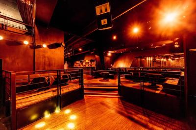 Salle de club vide avec éclairage tamisé, tables rondes, banquettes et escalier menant à une piste de danse.