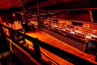 Salle de club vide avec éclairage tamisé, tables rondes, banquettes et escalier menant à une piste de danse.