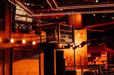Salle de club vide avec éclairage tamisé, tables rondes, banquettes et escalier menant à une piste de danse.