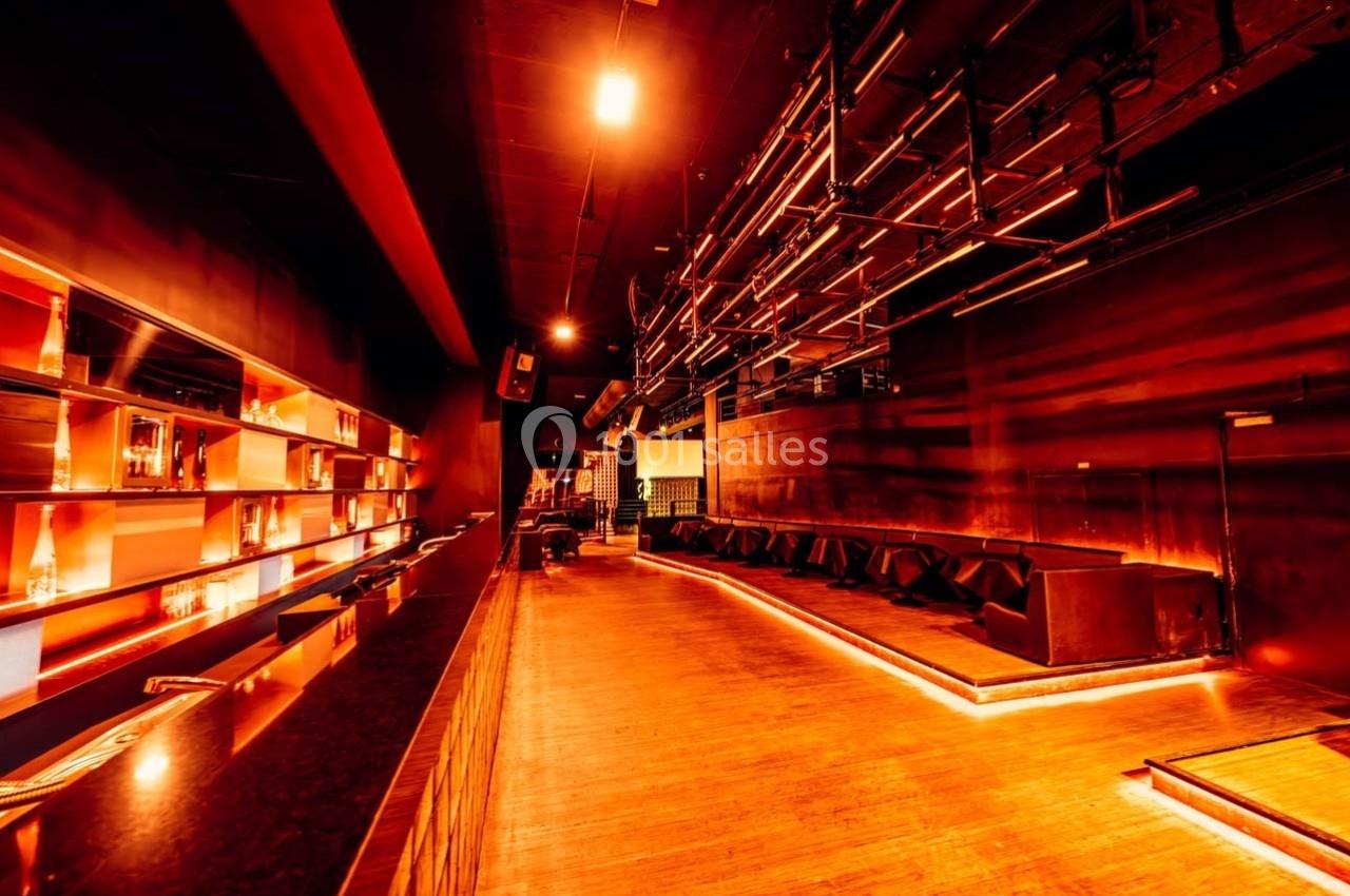 Salle de bar vide avec éclairage tamisé orange, comptoir à gauche et banquettes alignées à droite.