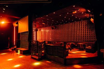 Salle de club vide avec éclairage tamisé, tables rondes, banquettes et escalier menant à une piste de danse.