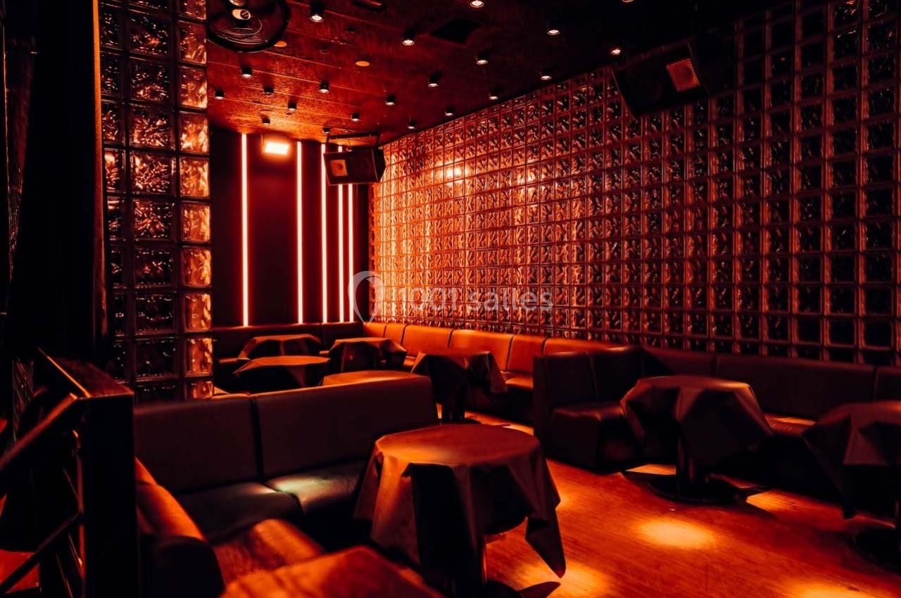 Salle sombre avec murs en briques de verre, éclairage rouge tamisé et tables recouvertes de nappes noires.