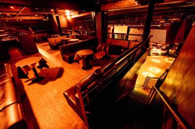 Salle de club vide avec éclairage tamisé, tables rondes, banquettes et escalier menant à une piste de danse.