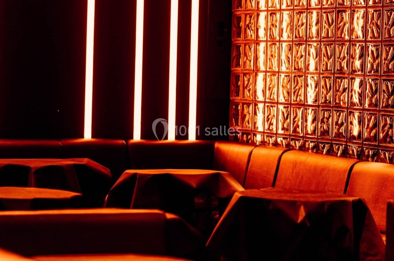 Banquettes et tables dans un espace intérieur sombre, éclairé par des néons rouges et un mur de briques de verre.