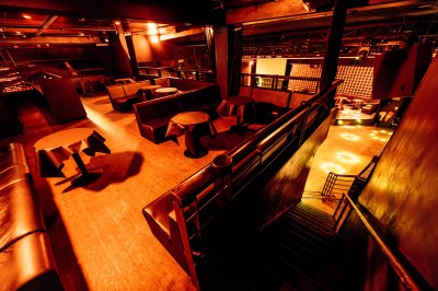 Salle de club vide avec éclairage tamisé, tables rondes, banquettes et escalier menant à une piste de danse.