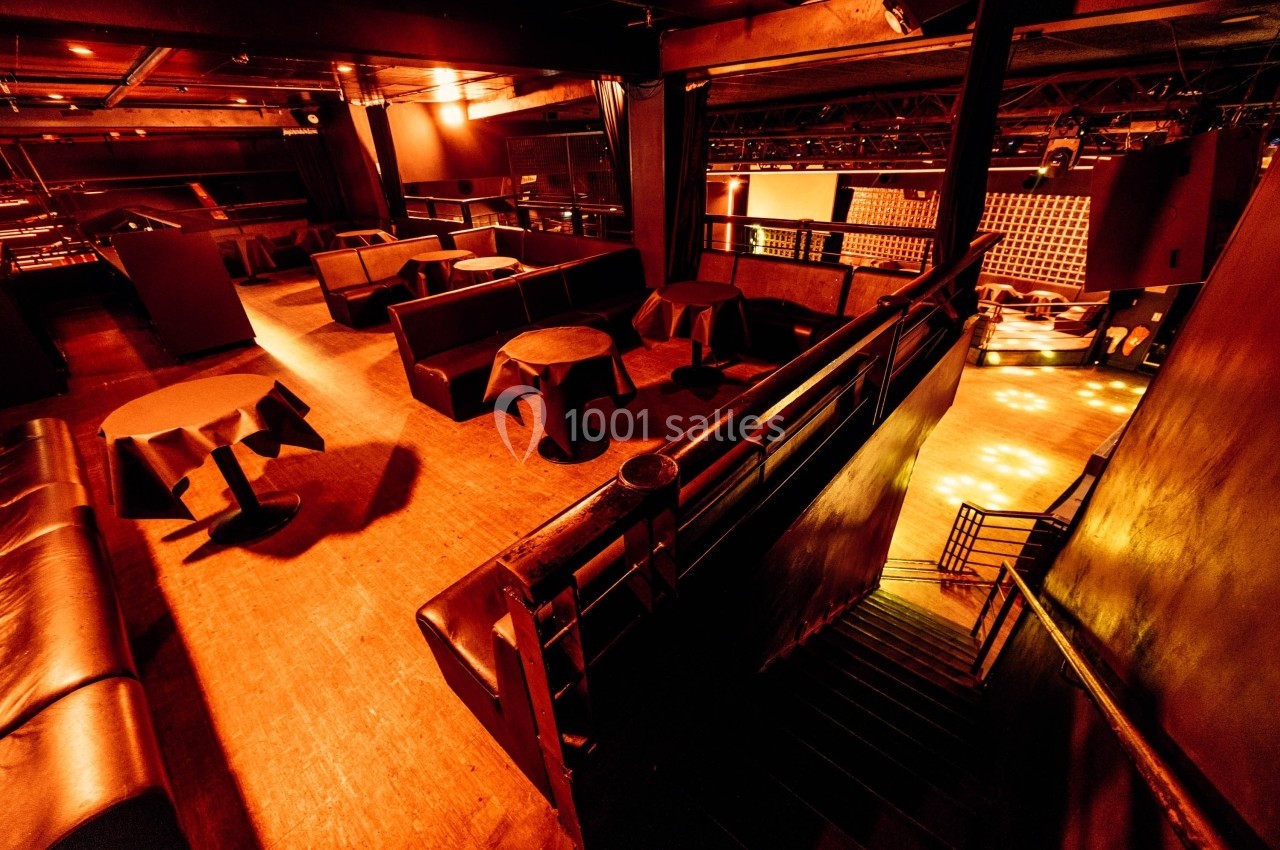 Salle de club vide avec éclairage tamisé, tables rondes, banquettes et escalier menant à une piste de danse.