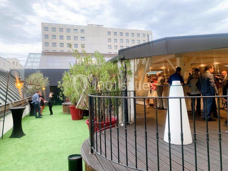 Terrasse extérieure avec pelouse synthétique, plantes en pot et personnes discutant près d'un espace couvert.