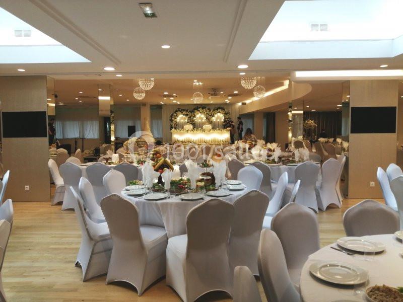 Salle de réception élégante avec tables rondes dressées, chaises blanches et décoration florale au fond.