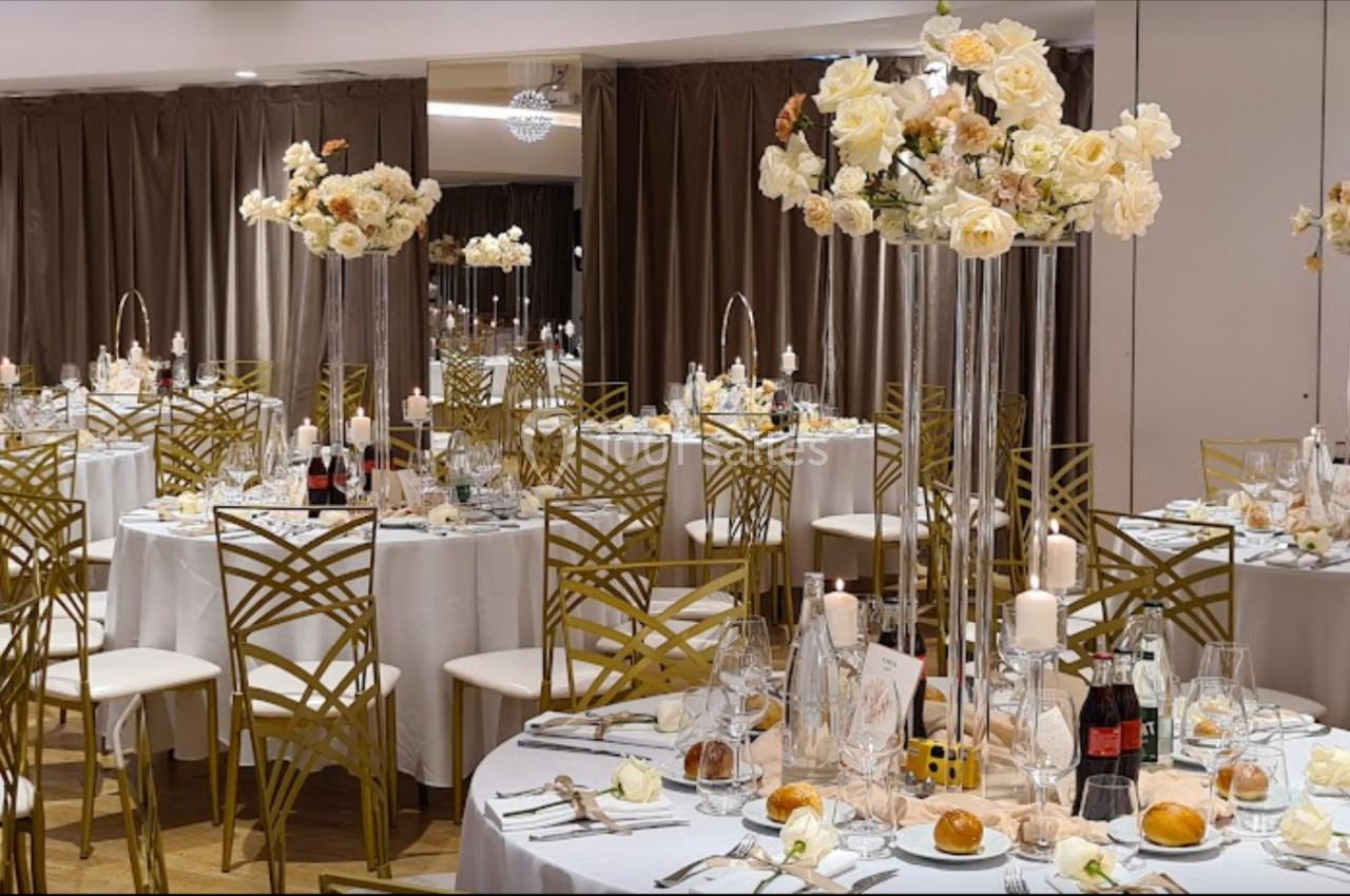 Salle de réception élégante avec tables rondes dressées, décorations florales blanches et chaises dorées.