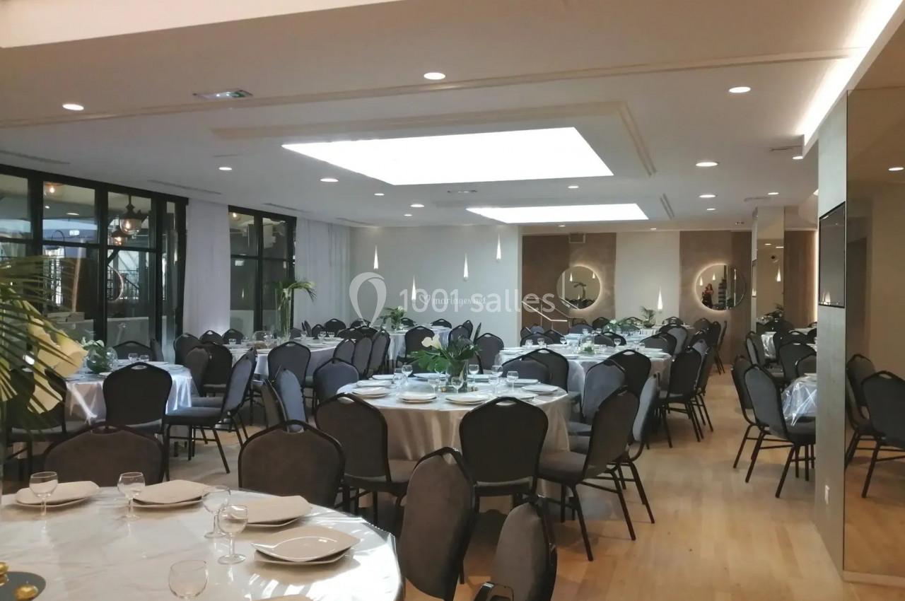 Salle de réception lumineuse avec tables rondes dressées, chaises noires et décoration sobre.