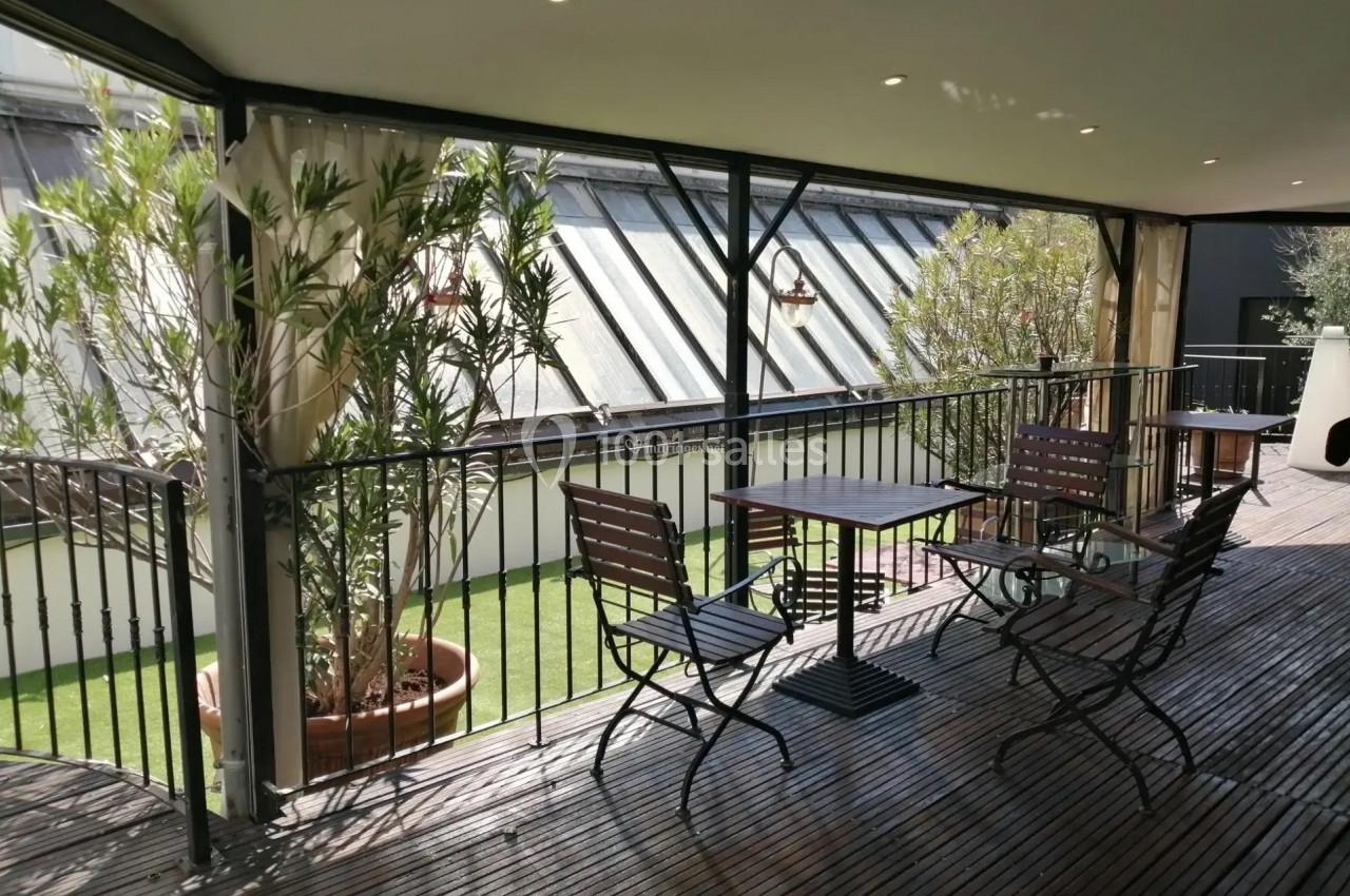 Terrasse couverte avec sol en bois, tables et chaises en métal, plantes en pot et vue sur un toit vitré.