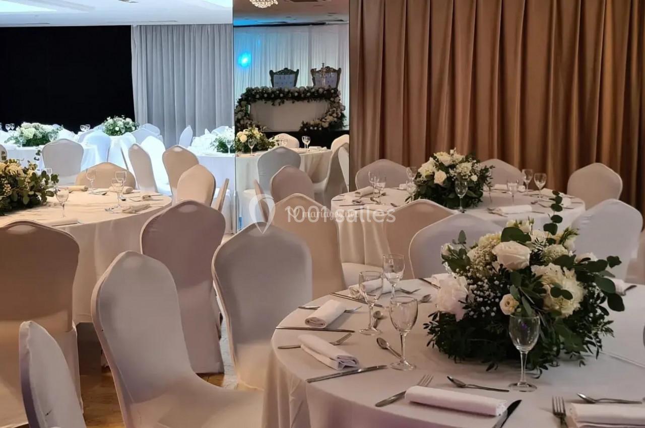 Salle de réception décorée avec des tables rondes, nappes blanches, chaises houssées et centres de table fleuris.
