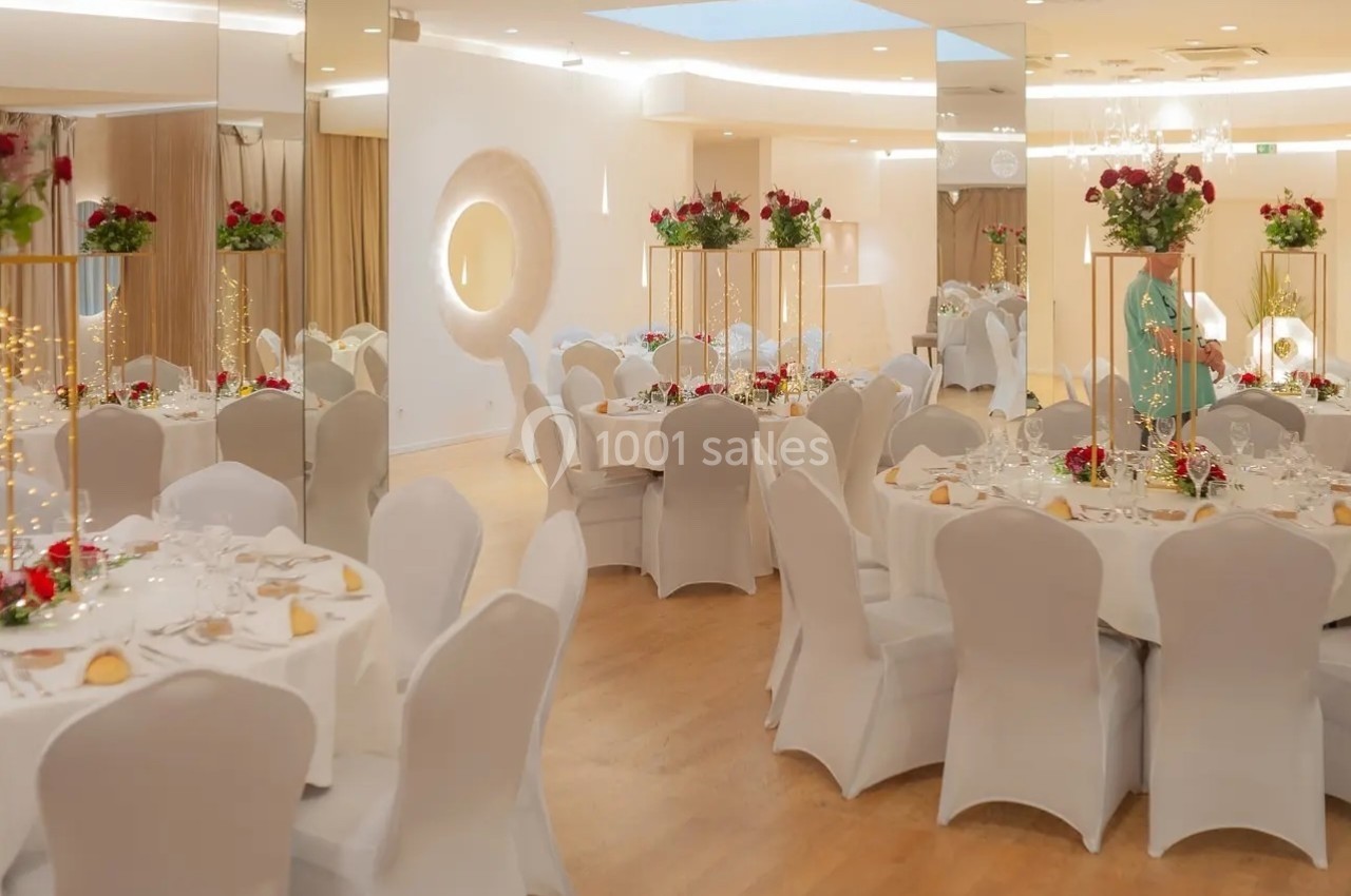 Salle de réception élégante avec tables rondes dressées, nappes blanches et décorations florales rouges et blanches.