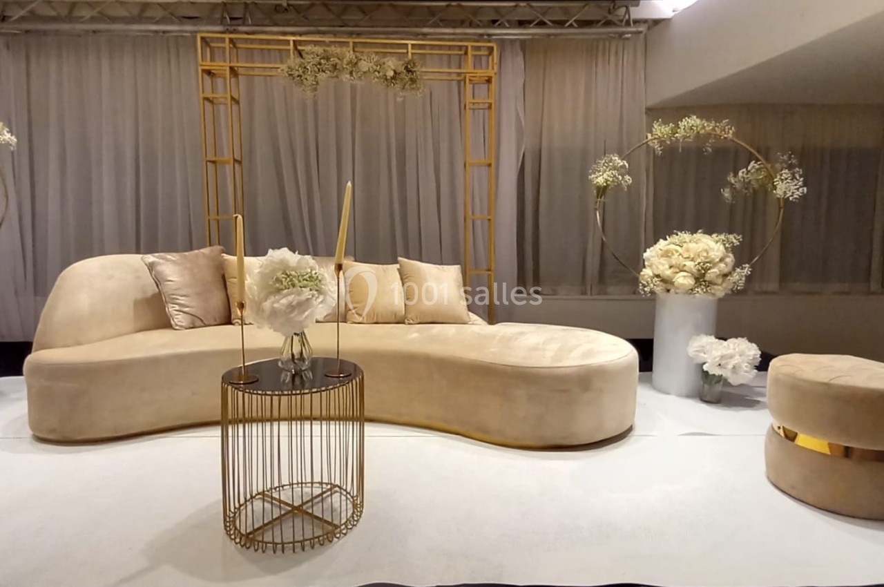 Salon élégant avec canapé beige incurvé, table basse dorée, décor floral blanc et arrière-plan de rideaux clairs.